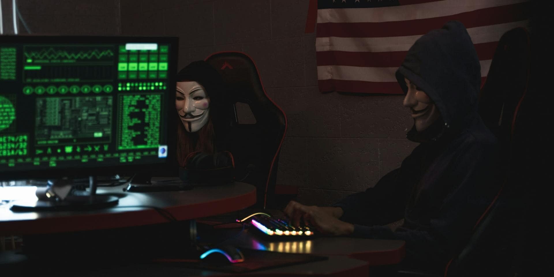 Anonymous hackers wearing Guy Fawkes masks hacking computers in a dimly lit room with the US flag in the background.
