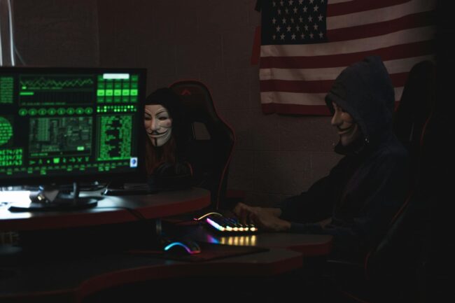Cyber Attackers Anonymous hackers wearing Guy Fawkes masks hacking computers in a dimly lit room with the US flag in the background.