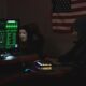 Anonymous hackers wearing Guy Fawkes masks hacking computers in a dimly lit room with the US flag in the background.