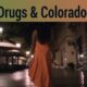 Woman walking alone at night in an urban area, illuminated by streetlights, representing themes of controversy surrounding sex work, drugs, and land disputes in Colorado.